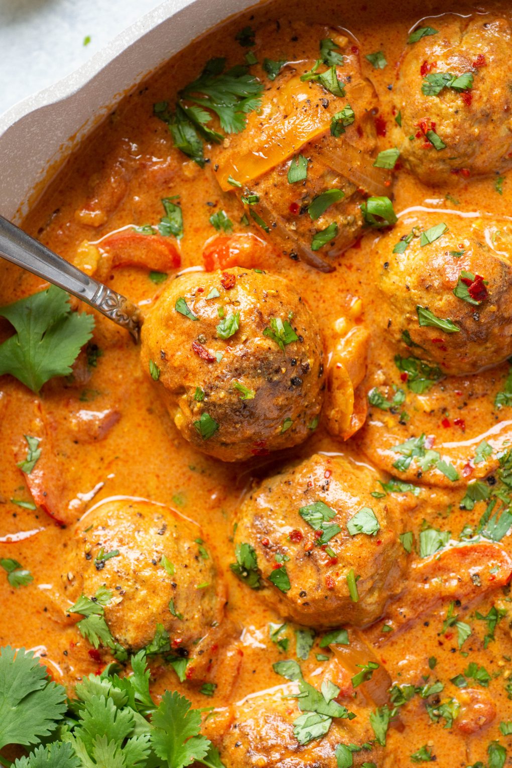 Overhead shot of a spoon holding a single paleo meatball in a skillet of meatballs and vibrant red curry sauce. 