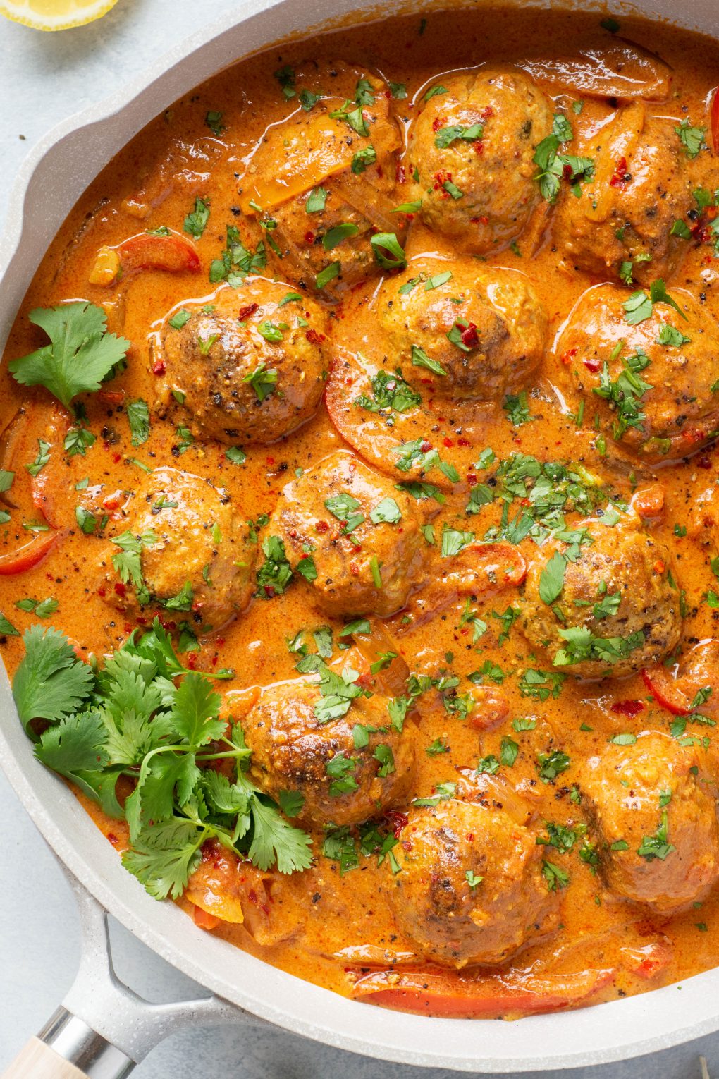 Overhead shot of a large white skillet filled with paleo meatballs in a vibrant red curry sauce. Topped with chopped fresh cilantro on a light colored background. 