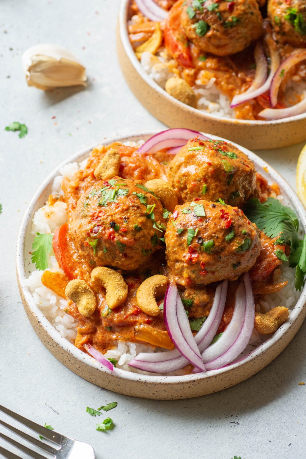 Side angle shot of a small white speckled plate on a light colored background filled with white rice topped with 3 meatballs in a vibrant red curry sauce. Topped with chopped cilantro, thin slices of red onion, and curried cashews. 