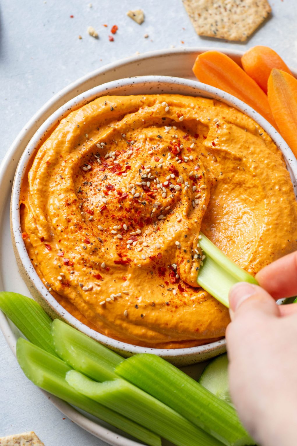 Side angle shot of a small speckled white bowl filled with bright orange roasted red pepper tahini dip. Dip is garnished with sesame seeds and chili flakes, and a hand on the bottom right of the frame is dipping a celery stick into the dip. On a plate with carrots, sliced cucumber, and celery sticks.