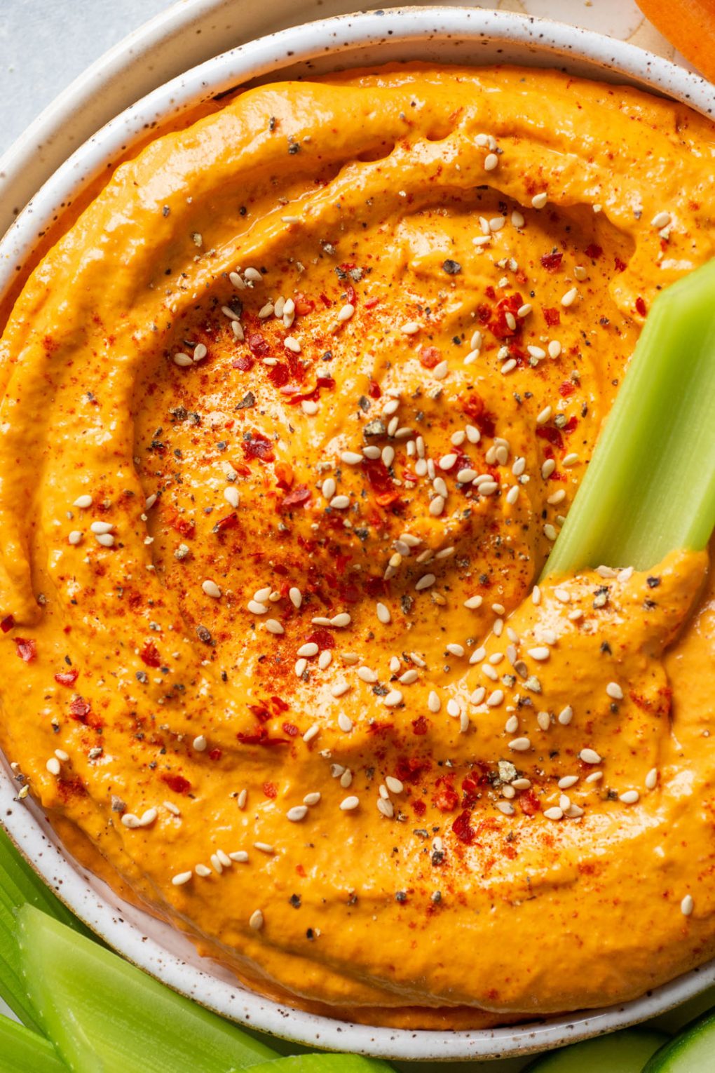 Close up overhead shot of a small speckled white bowl filled with bright orange roasted red pepper dip. Dip is garnished with sesame seeds and chili flakes. On a plate with carrots, sliced cucumber, and celery sticks.