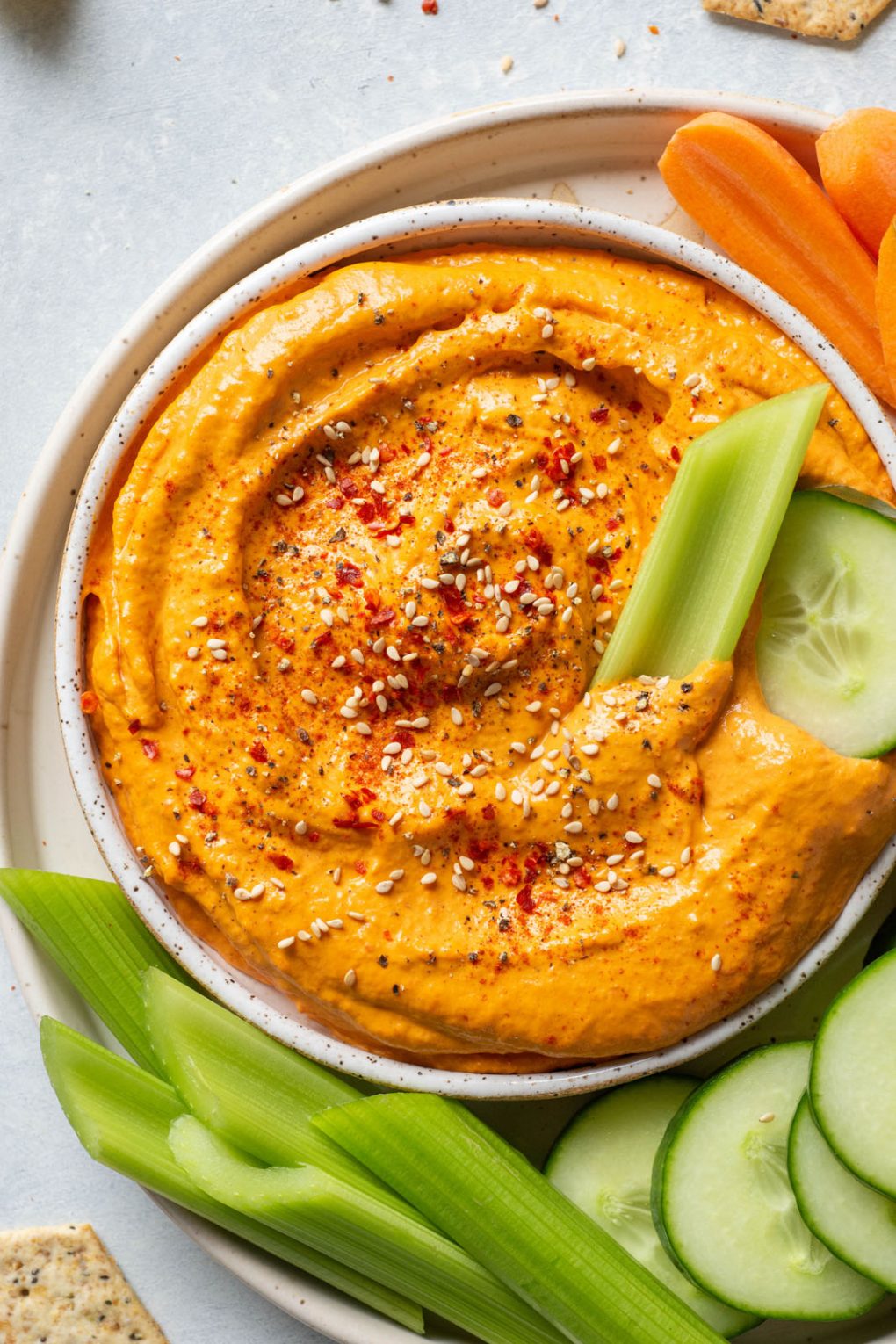 Overhead shot of a small speckled white bowl filled with bright orange roasted red pepper tahini dip. Dip is garnished with sesame seeds and chili flakes. On a plate with carrots, sliced cucumber, and celery sticks.