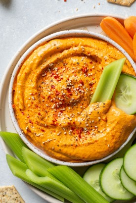 Overhead shot of a small speckled white bowl filled with bright orange roasted red pepper tahini dip. Dip is garnished with sesame seeds and chili flakes. On a plate with carrots, sliced cucumber, and celery sticks.