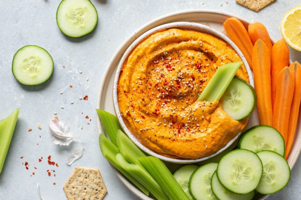 Overhead landscape shot of a small speckled white bowl filled with bright orange roasted red pepper tahini dip. Dip is garnished with sesame seeds and chili flakes. On a plate with carrots, sliced cucumber, and celery sticks.