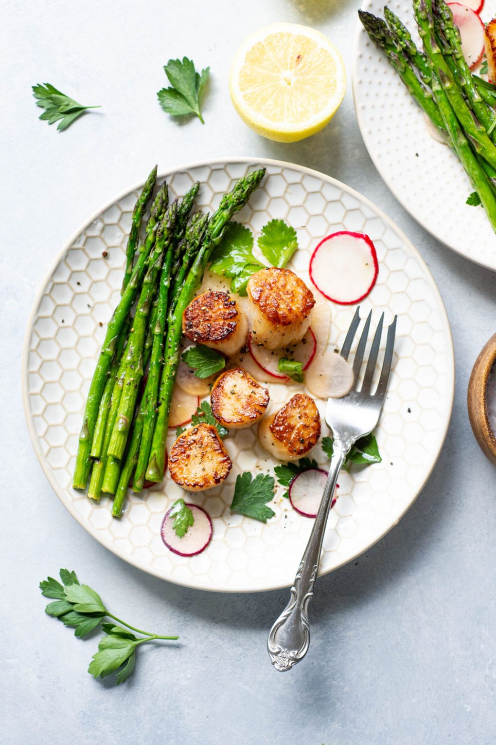 Overhead shot of a white plate with seared scallops over a thinly sliced radish salad, next to a bundle of bright green asparagus with a silver fork on the plate. On a light background surrounded by a cut lemon and a scatter of fresh herbs.