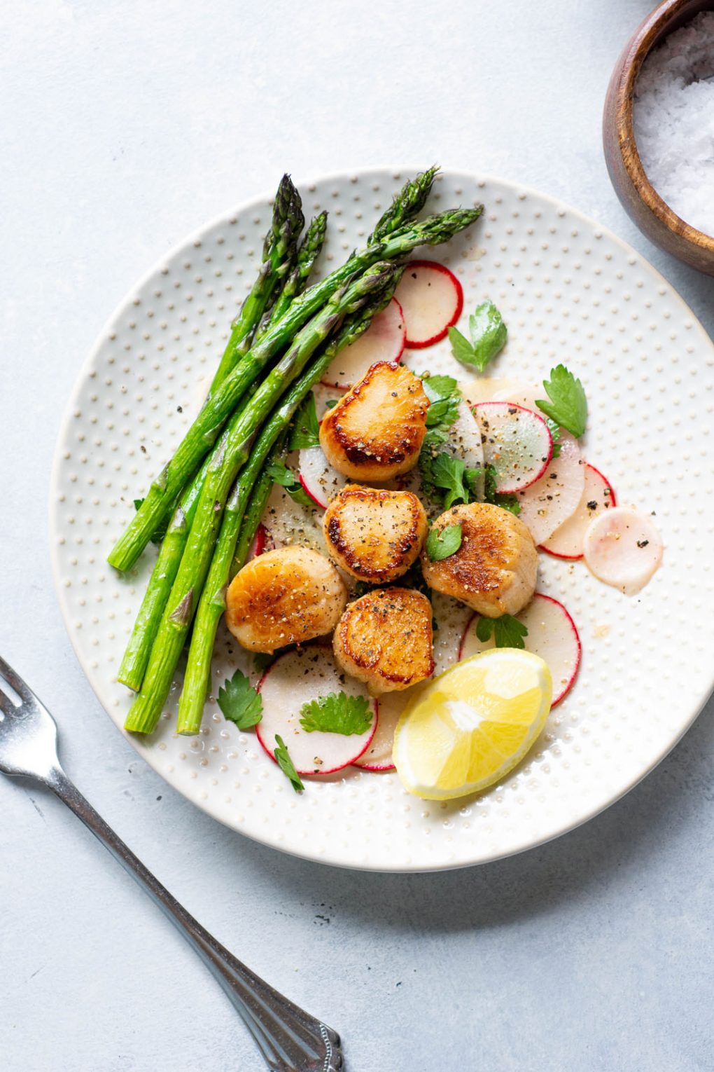 Overhead shot of a white plate with seared scallops over a thinly sliced radish salad, next to a bundle of bright green asparagus. On a light background next to a wooden bowl of flaky sea salt and a silver fork.