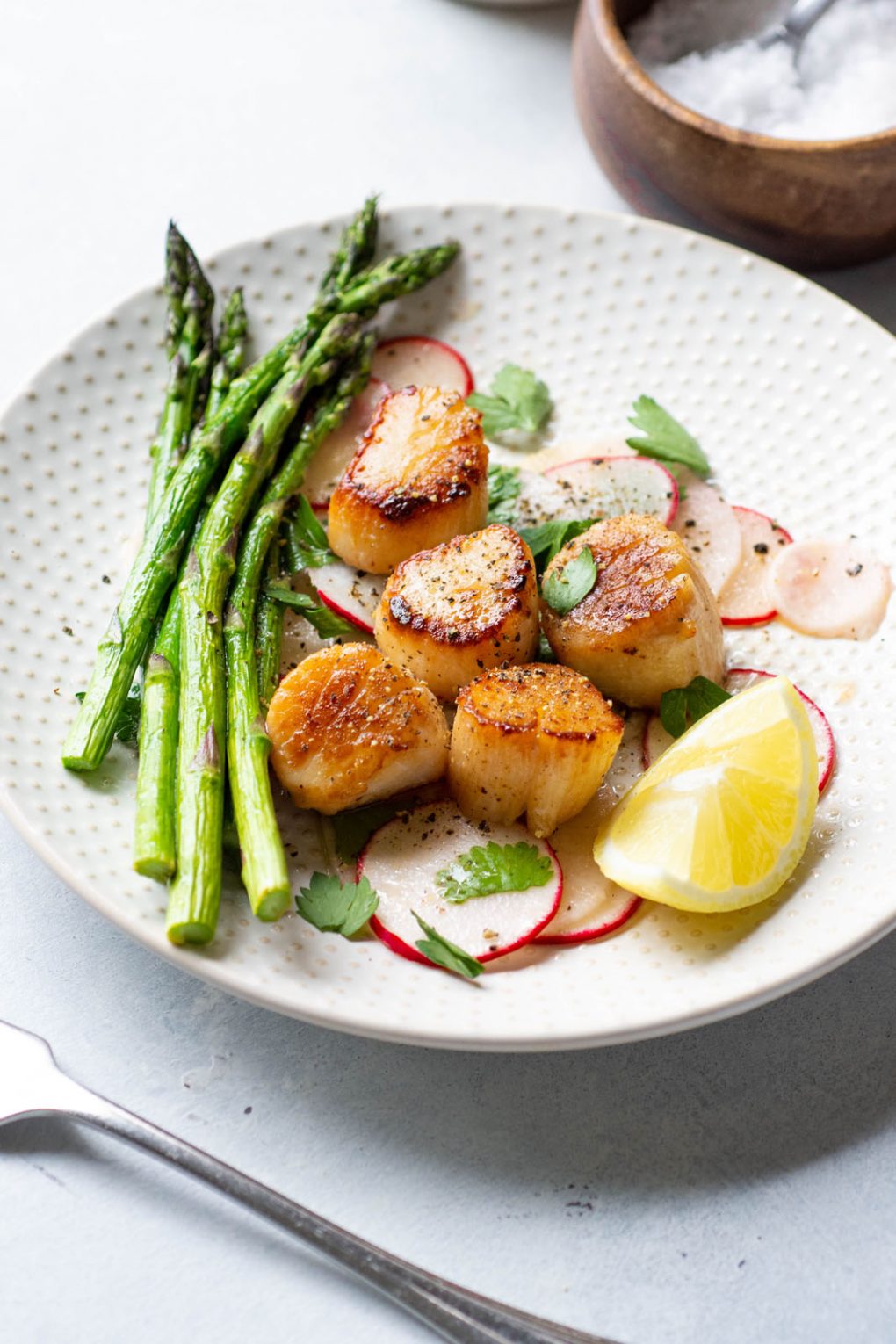 Side angle shot of a white plate with seared scallops over a thinly sliced radish salad, next to a bundle of bright green asparagus. On a light background next to a wooden bowl of flaky sea salt and a silver fork.