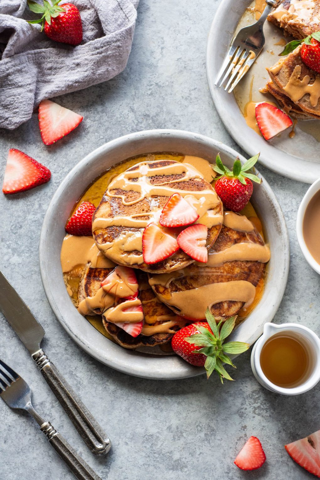 Overhead view of a plate of pancakes that are drizzled with almond butter and fresh strawberries. On a grey plate and light grey background.