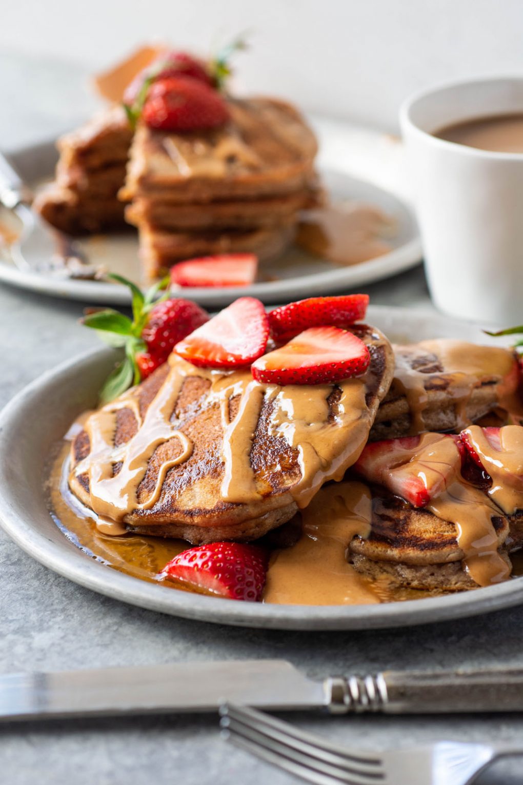 Side angle view of a plate of pancakes that are drizzled with almond butter and fresh strawberries. On a grey plate and light grey background.