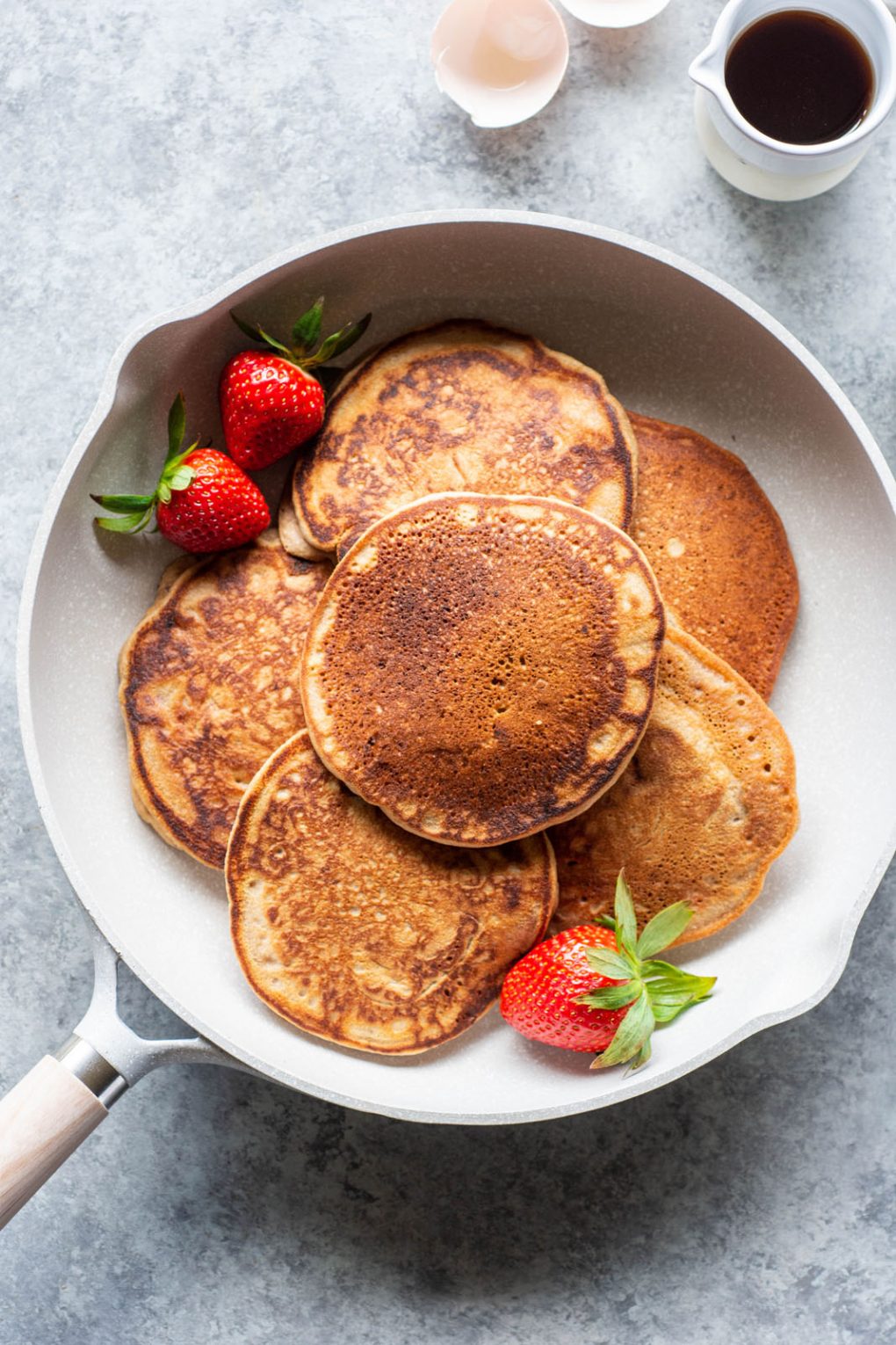 Overhead shot of a white skillet with perfectly golden brown pancakes surrounded by fresh strawberries. On a light grey background.