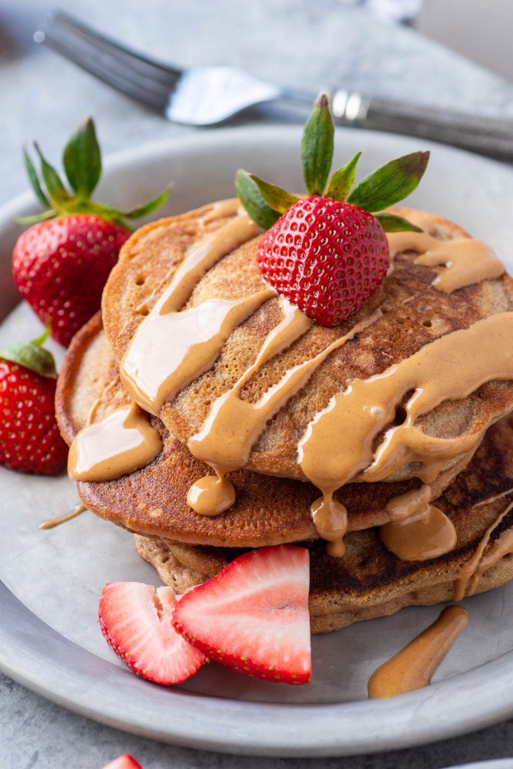 Close up side angle shot of a stack of pancakes drizzled with almond butter and topped with fresh cut strawberries. 