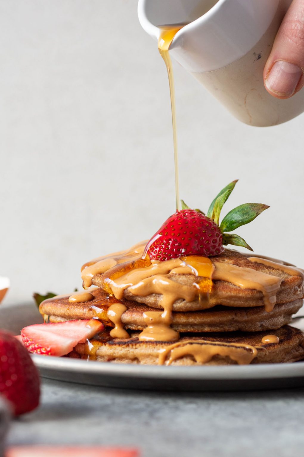 Side angle view of a stack of pancakes being drizzled with maple syrup. Topped with fresh strawberries on a light grey background.