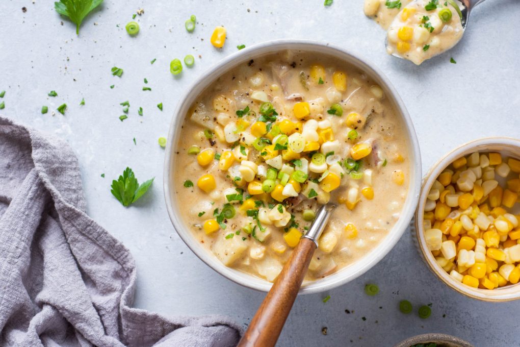 Overhead shot of a white bowl of creamy corn chowder topped with sliced green onions and chopped parsley. On a white background with an angled wooden soup spoon in the bowl. Surrounded by a smaller bowl filled with corn, and scattered pieces of herbs and corn kernals. 