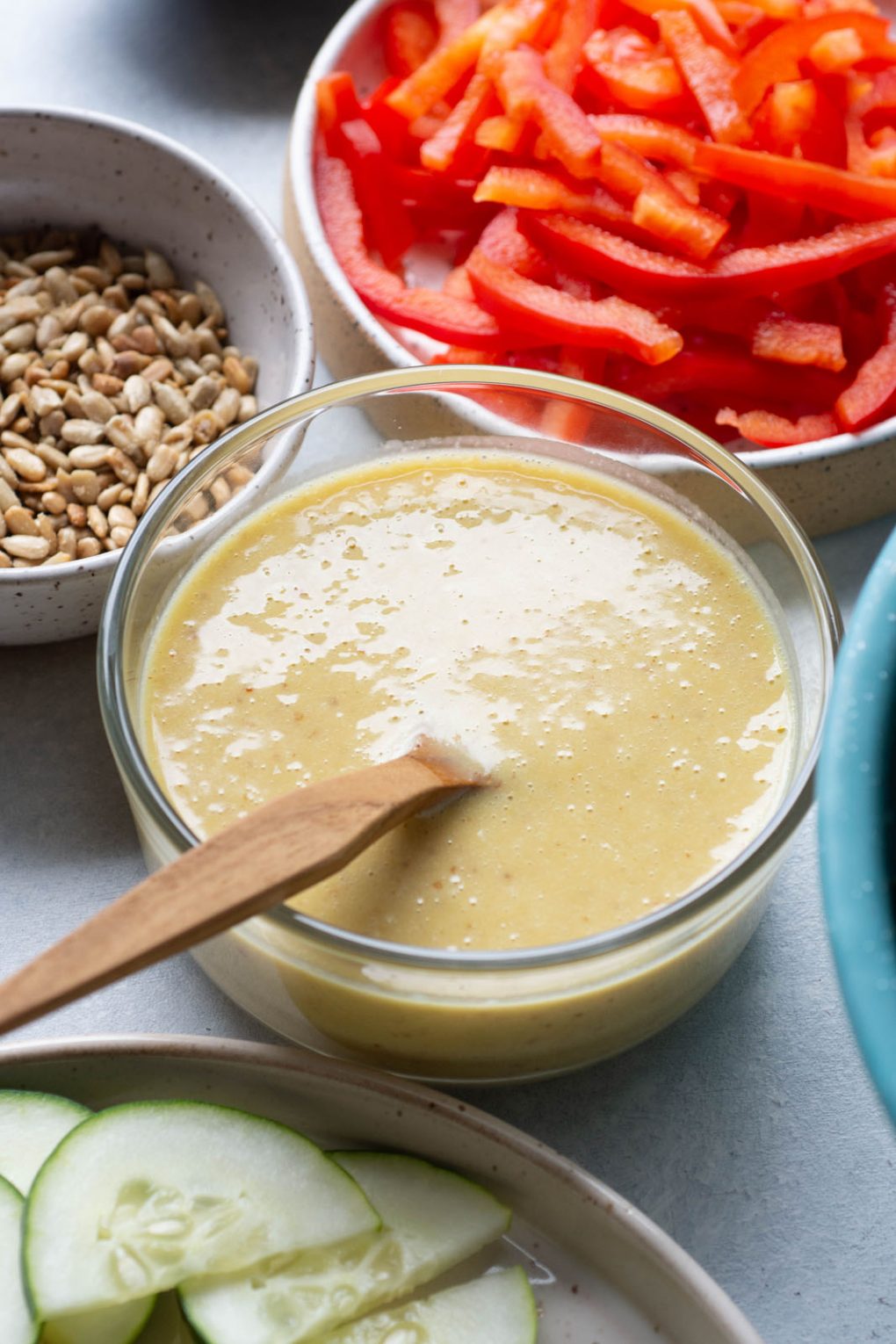 Side angle shot of a small glass pyrex of honey mustard dressing with a wooden spoon. On a light background surrounded by several ingredient bowls.