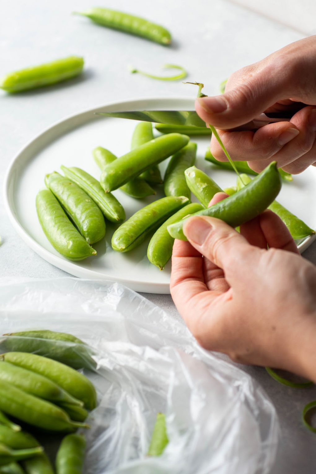 Side angle view of two hands pulling the string off of a sugar snap pea with a paring knife - on a light colored background next to a plate filled with sugar snap peas and a plastic produce bag holding the peas that have yet to be trimmed.