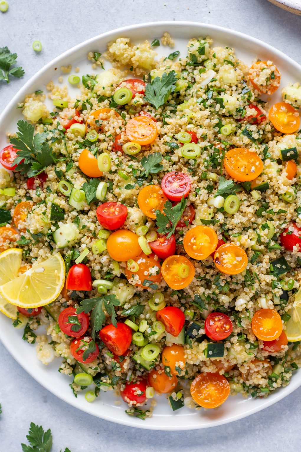 Overhead shot of a large colorful plate of quinoa tabbouleh with red and orange cherry tomatoes, cucumber, parsley, and green onions. Garnished with a few lemon wedges on a light colored background surrounded by some scattered herbs and cherry tomatoes. 