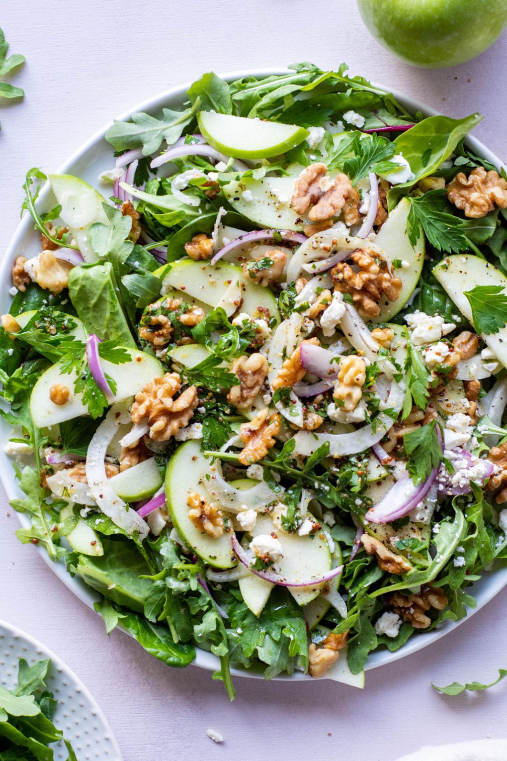 Large green salad on a round plate and a light colored background. Salad greens layered with thinly sliced green onion, thinly shaved fresh fennel, sliced red onion, walnuts, and torn parsley leaves. Drizzled with a mustardy dressing.
