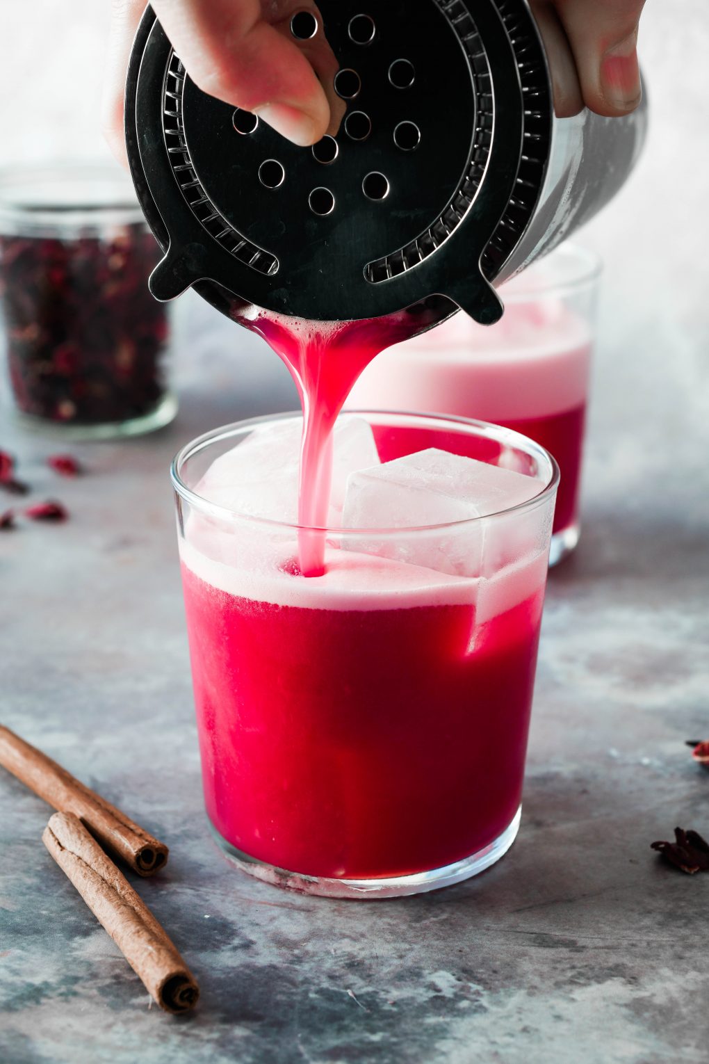 Close up shot of straining the spiced hibiscus sour cocktail into a glass filled with ice. On a dark grey background with 2 cinnamon sticks angled next to the glass.