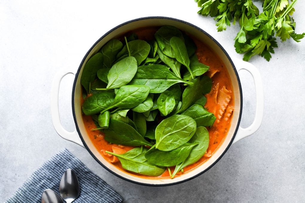 Horizontal overhead shot of a large dutch oven filled with lasagna soup, piled high with raw baby spinach.
