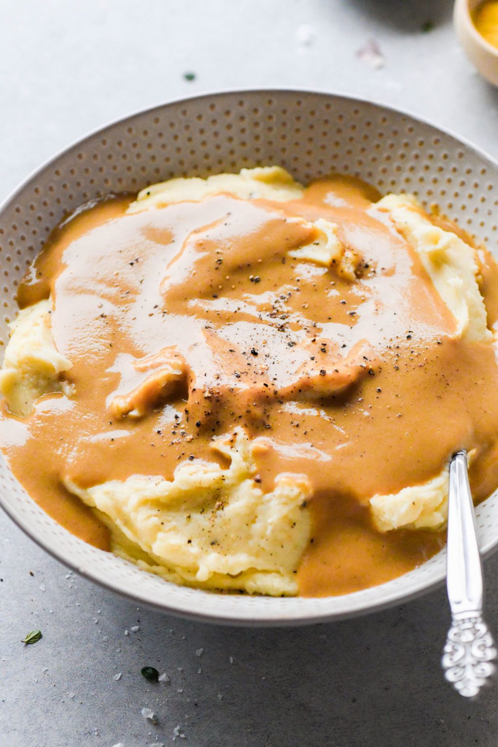 45 degree angle shot of super easy gluten free gravy draped over mashed potatoes, and topped with some cracked black pepper, with a spoon tucked into the mashed potatoes. On a light grey background. 