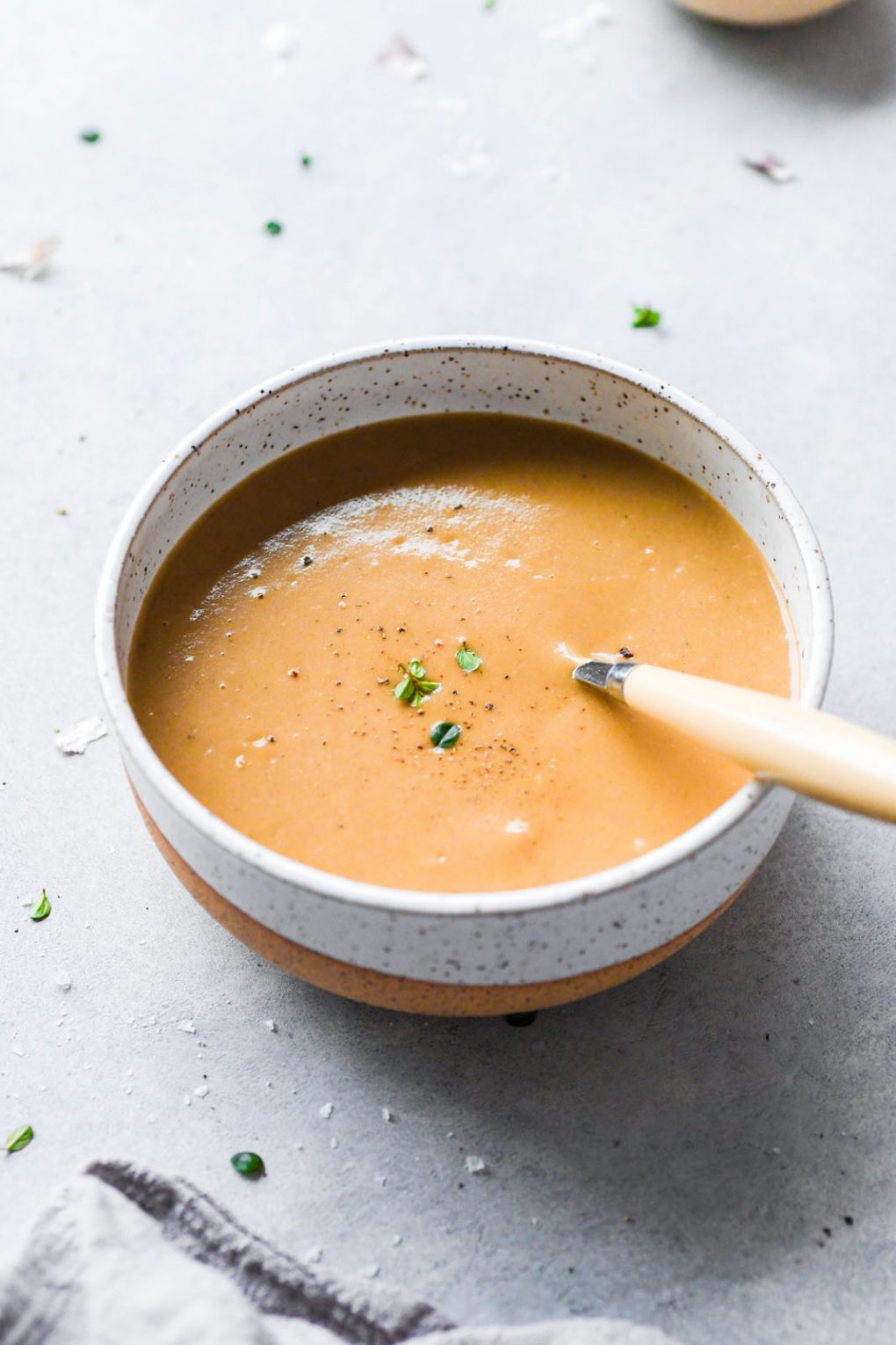 Side angle shot of a bowl of super easy gluten free gravy with a spoon resting inside the bowl. On a light grey background scattered with thyme leaves and flaky sea salt. 
