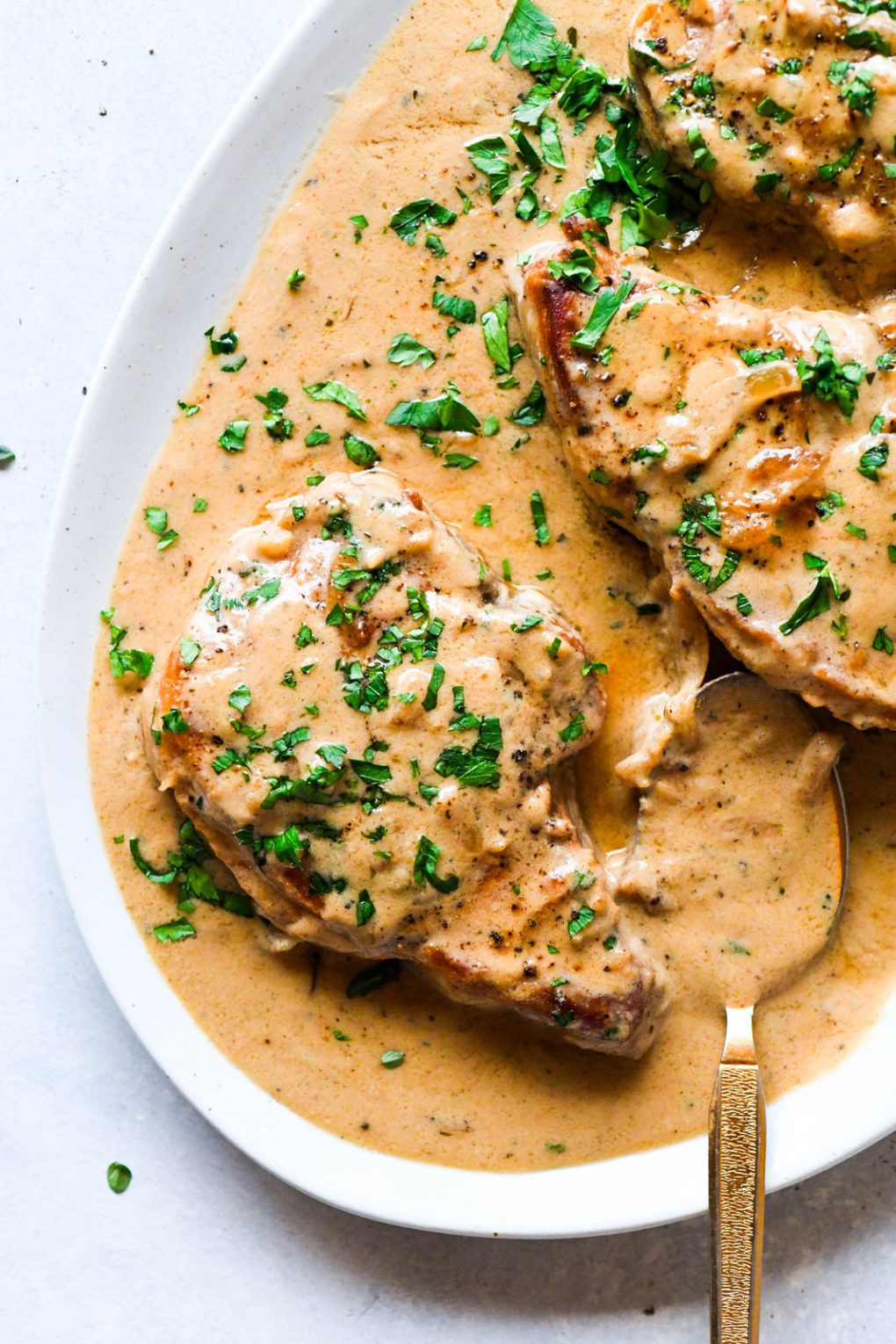 Overhead shot of a platter with smothered pork chops in plenty of creamy cashew gravy, with a spoon dipped into the gravy. Topped with chopped parsley on a light colored background. 