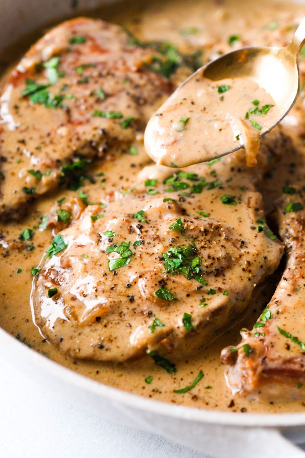 Close up shot of a spoon drizzling super creamy cashew gravy over a golden brown pork chop in a skillet topped with chopped parsley. 
