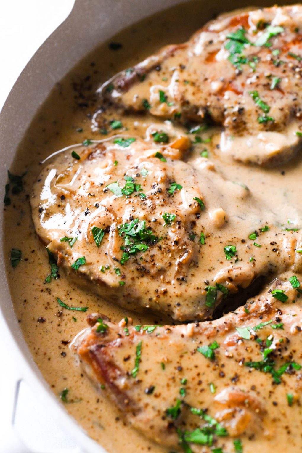 Side angle shot of cooked pork chops in a creamy gravy, topped with chopped parsley. 