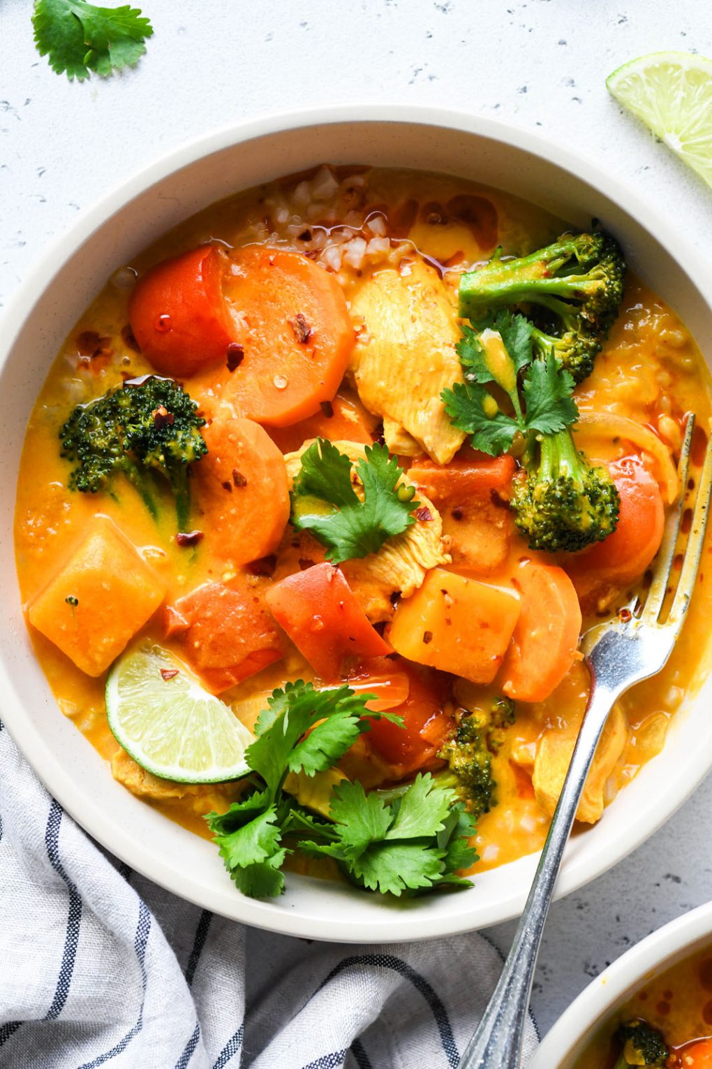 Close up overhead shot of a bright orange bowl of pumpkin curry. Brothy curry with sliced carrots, red pepper pieces, broccoli, chicken, and garnished with lime wedges and fresh cilantro. 