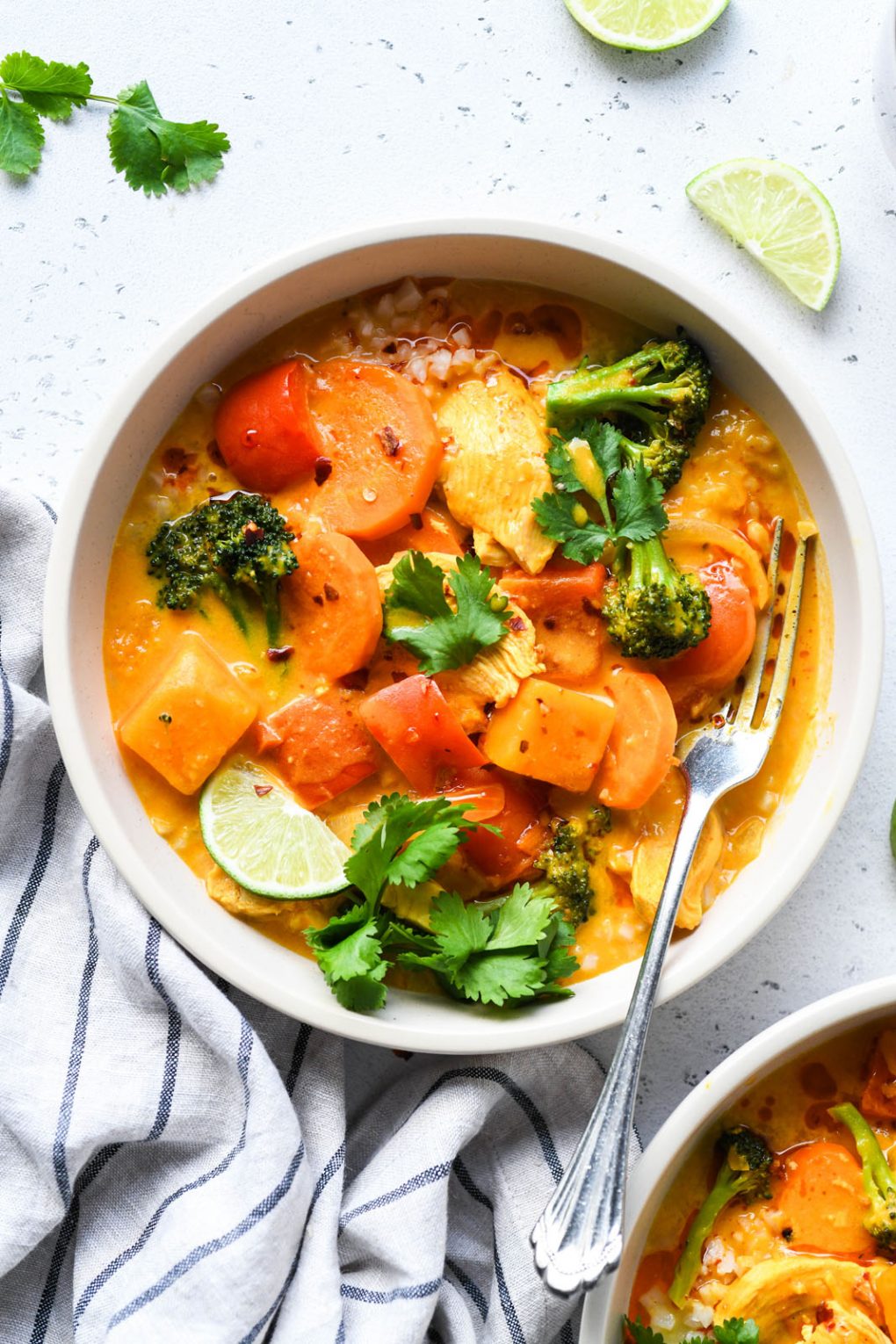 Close up overhead shot of a bright orange bowl of pumpkin curry. Brothy curry with sliced carrots, red pepper pieces, broccoli, chicken, and garnished with lime wedges and fresh cilantro. On a light colored background next to a striped napkin surrounding the bowl. 