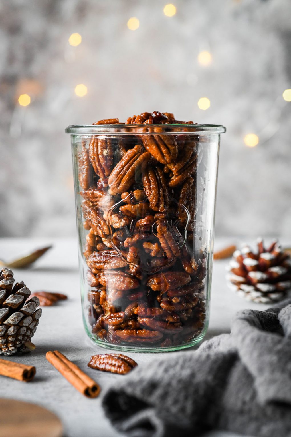 Close up shot of a tall weck jar filled with candied pecans. With a dark grey background, surrounded by a few festive pinecones and cinnamon stick.