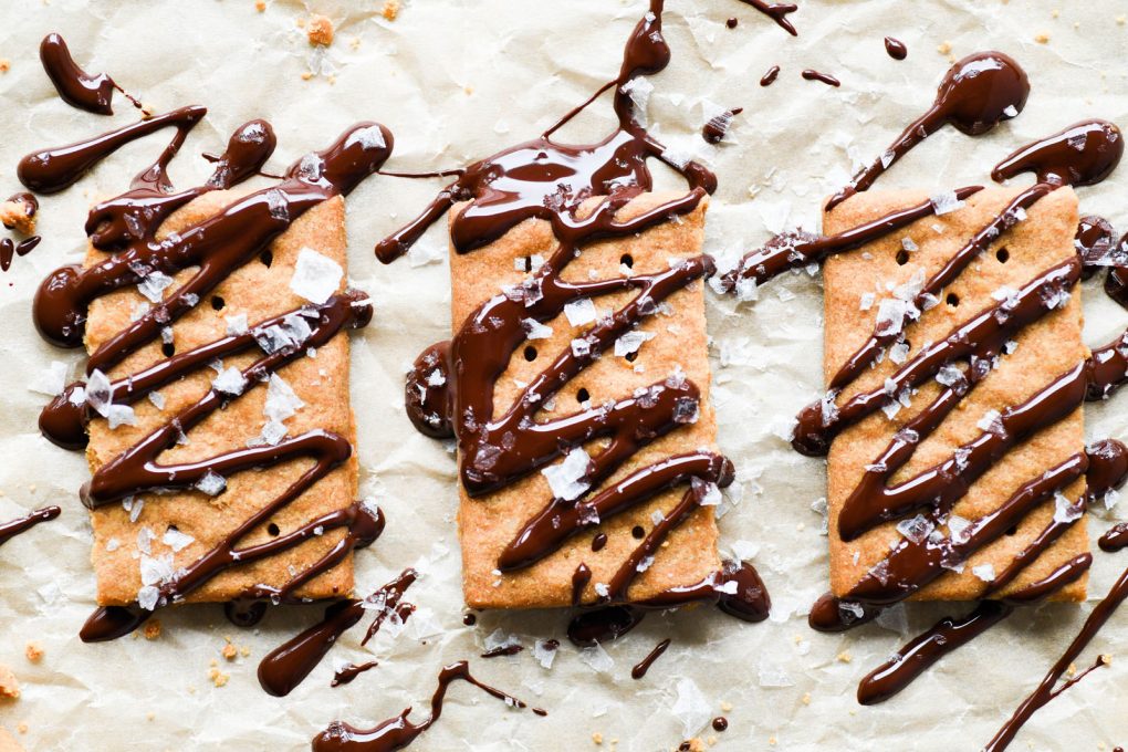 Overhead view of graham crackers drizzled with melted chocolate and topped with flaky sea salt, on light brown parchment paper.