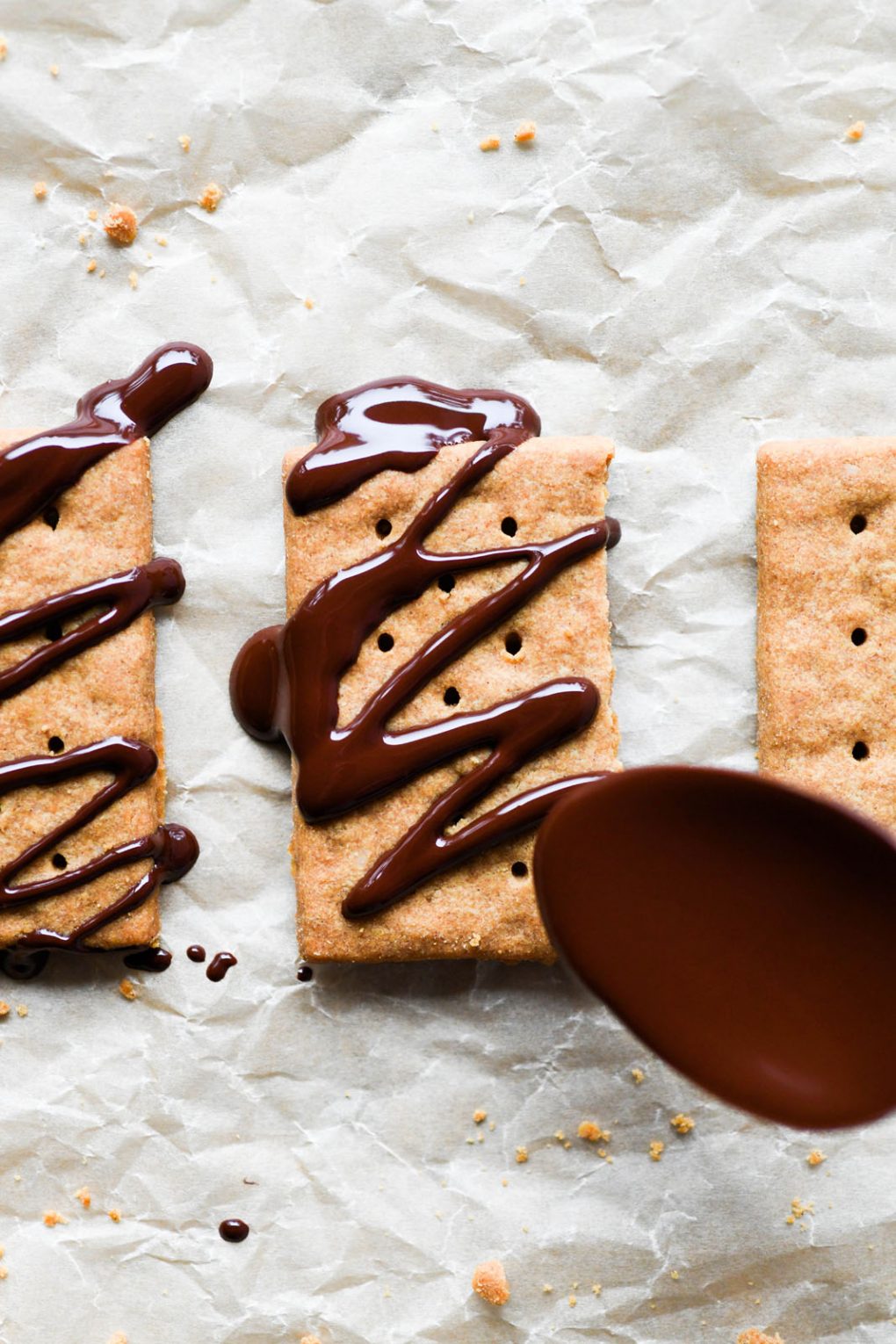 Overhead view of a spoon drizzling melted chocolate over graham crackers, on light brown parchment paper.