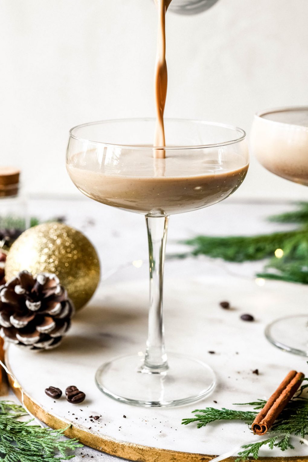 Straight on shot of pouring the eggnog espresso martini into a couple glass. Coupe glass is next to a pine cone, some scattered coffee beans, and a gold sparkly christmas tree ornament. 