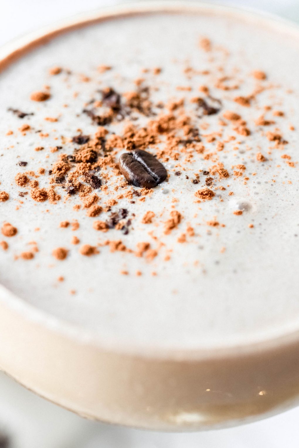 Super close up shot of the surface of a creamy eggnog espresso martini - In frame is creamy white foam scattered with the cocktail garnish - cinnamon, ground coffee, and a coffee bean. 