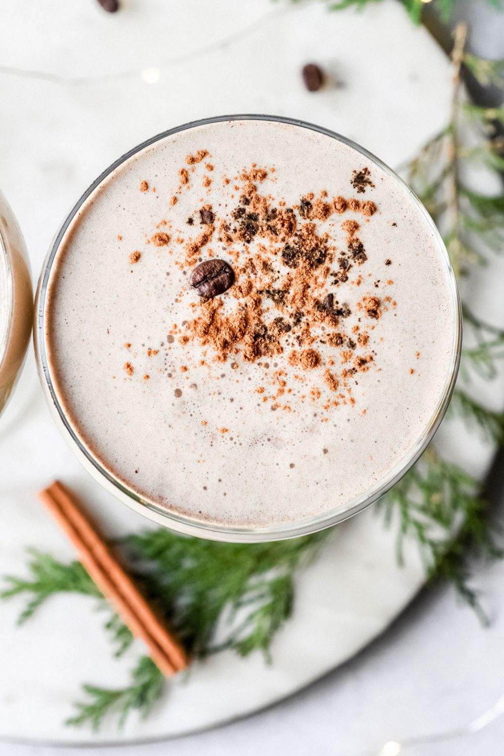 Overhead shot of a creamy eggnog espresso martini in a coupe glass. Topped with a dash of cinnamon and a coffee bean. On a white background surrounded by cedar, and pine cones.