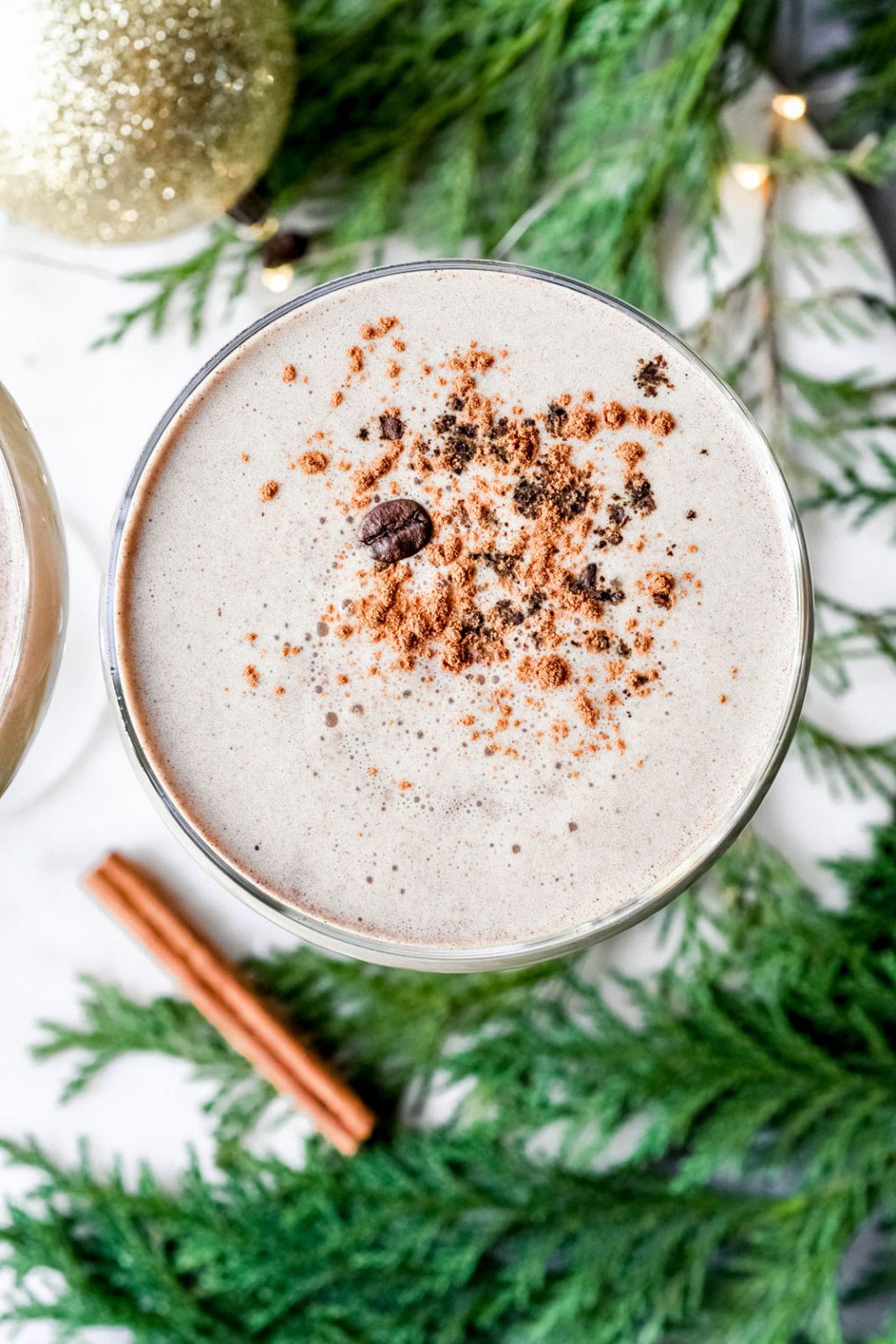 Close up overhead shot of a creamy eggnog espresso martini in a coupe glass. Topped with a dash of cinnamon and a coffee bean. On a white background surrounded by cedar, pine cones and tiny little string lights. 