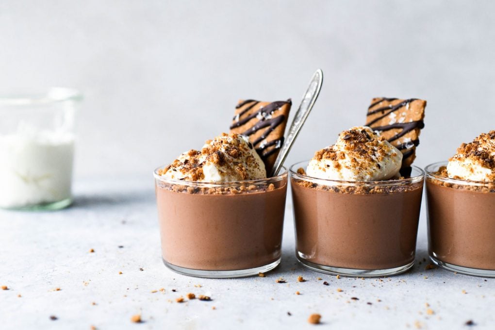 Straight on shot of three vegan mousse cups in a row on a light colored background topped with whipped cream and chocolate drizzled graham crackers. 