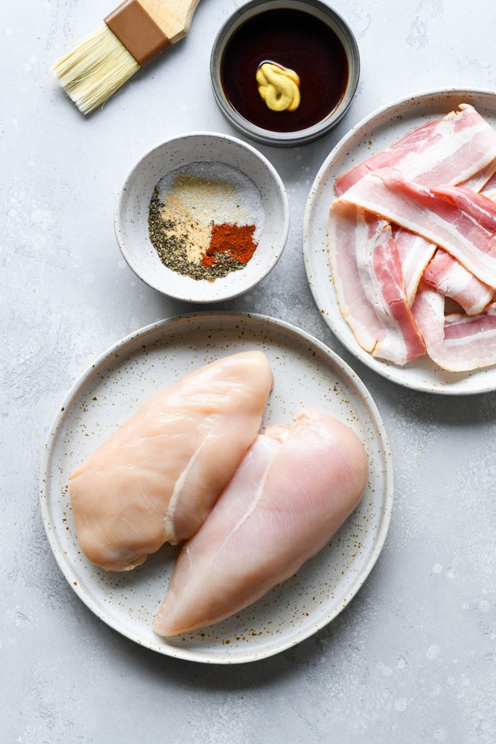 An image of the ingredients for whole30 bacon wrapped chicken. Two large chicken breasts on a white speckled plate, a small pile of bacon, spices in a little white bowl, and a tiny dish with coconut aminos and dijon mustard. On a light colored background.