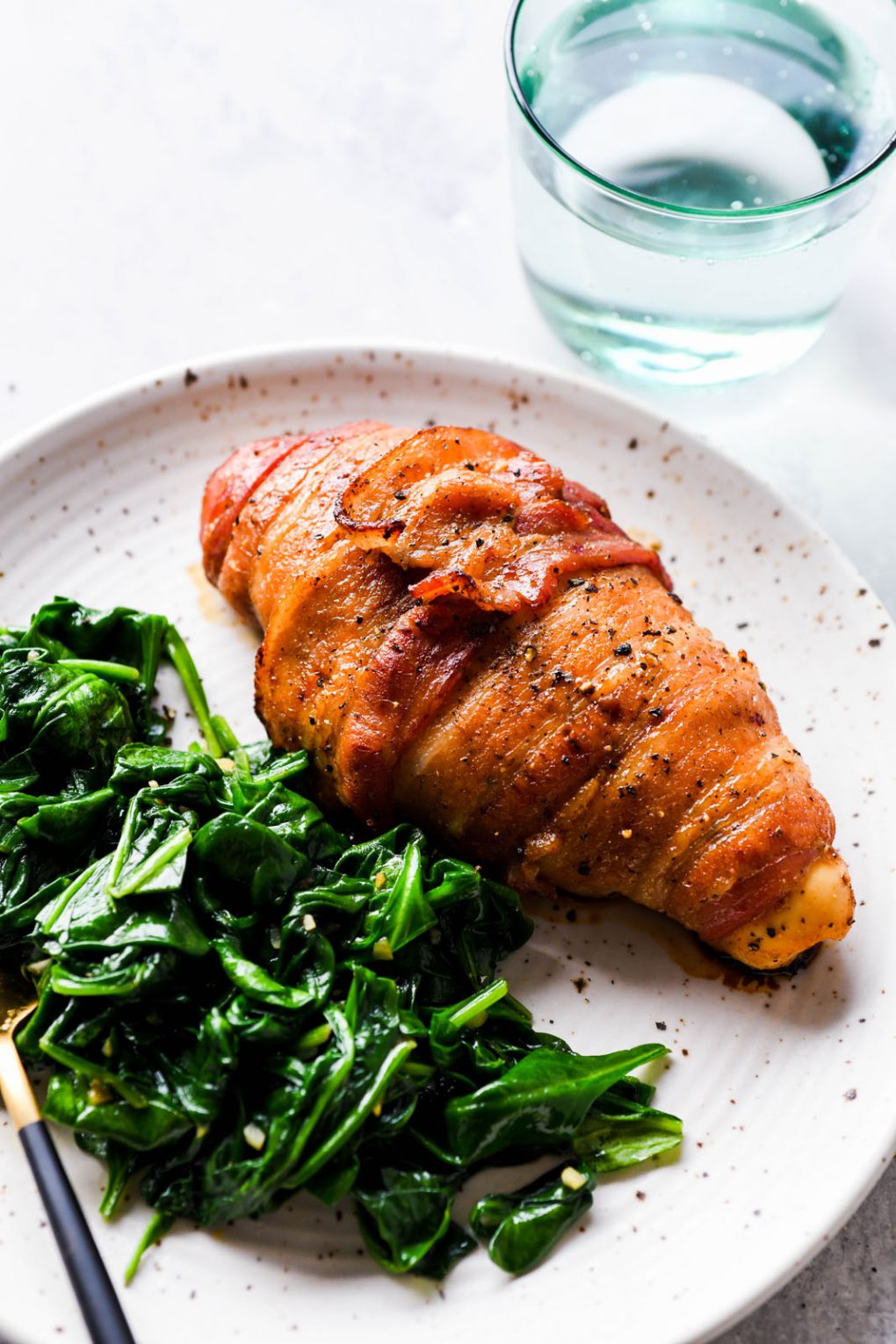 45 degree angle shot of a golden brown baked bacon wrapped chicken breast on a white speckled plate, next to a generous portion of wilted spinach with garlic. On a light colored background next to a light green glass with sparkling water. 