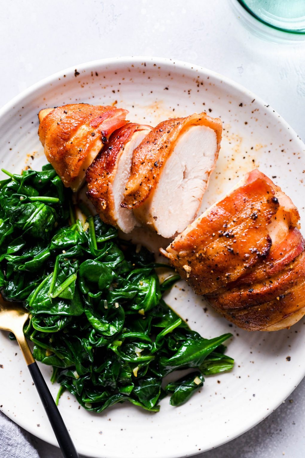 Close up image of a glistening bacon wrapped chicken breast cut into slices, on a white speckled plate next to some wilted garlic spinach, with a gold and black fork tucked onto the plate at an angle. 
