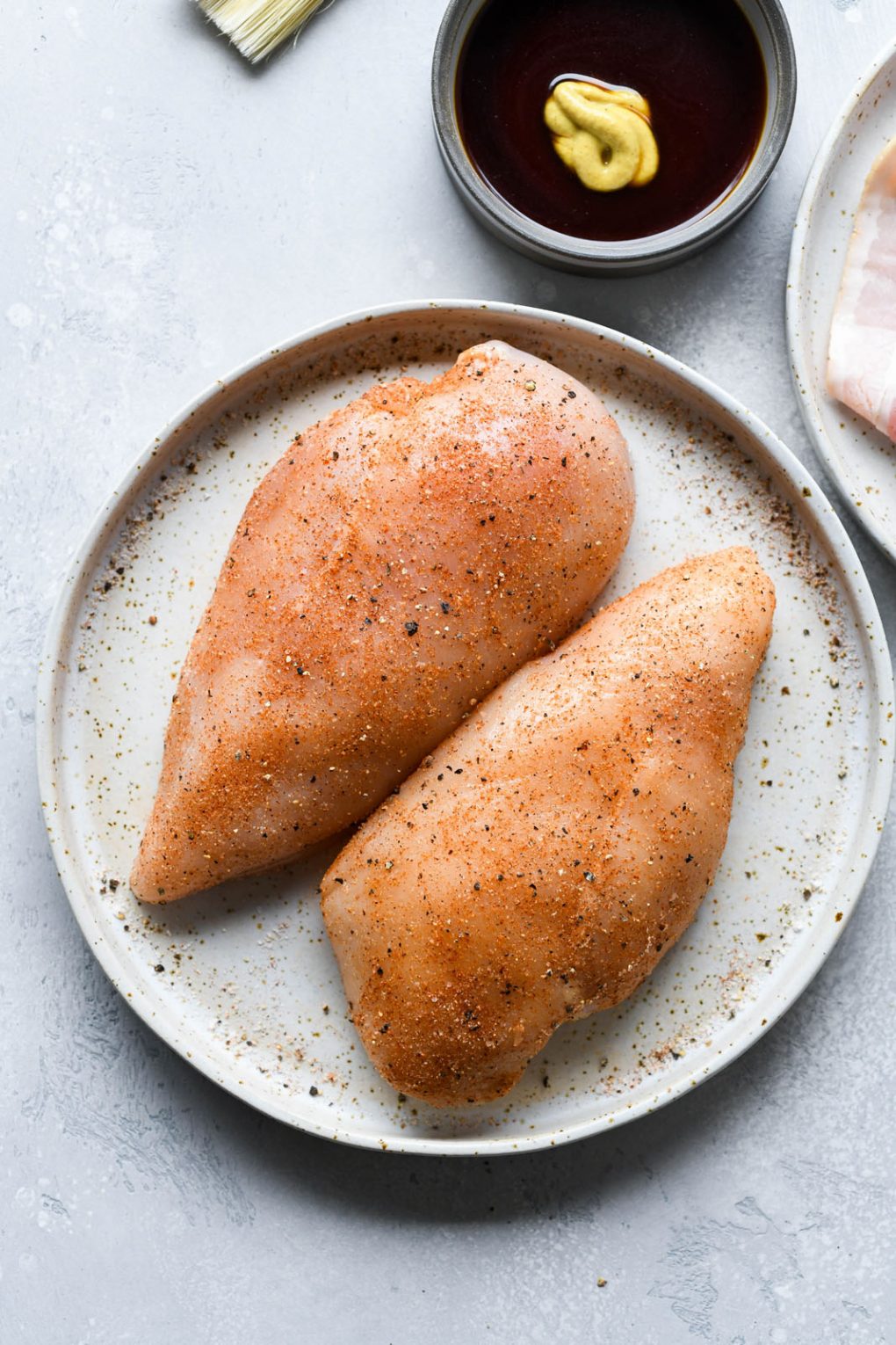 Image of two large raw chicken breasts coated in a spice mix, on a light colored plate - on a light background.