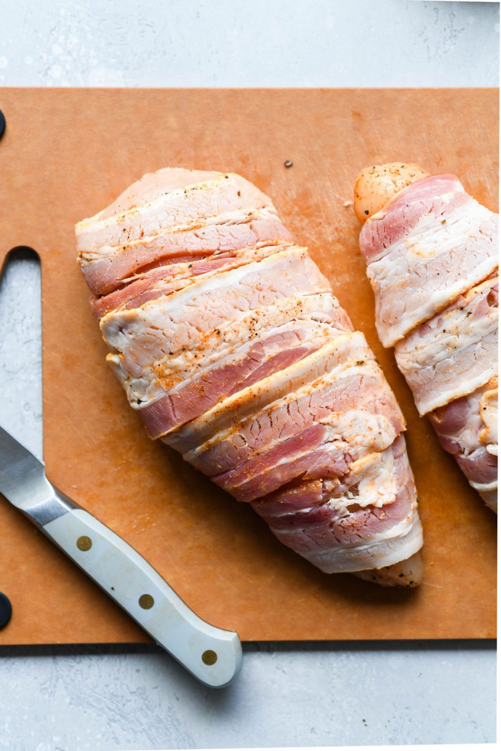 Close up image of two large chicken breasts wrapped in bacon, on a light brown cutting board.