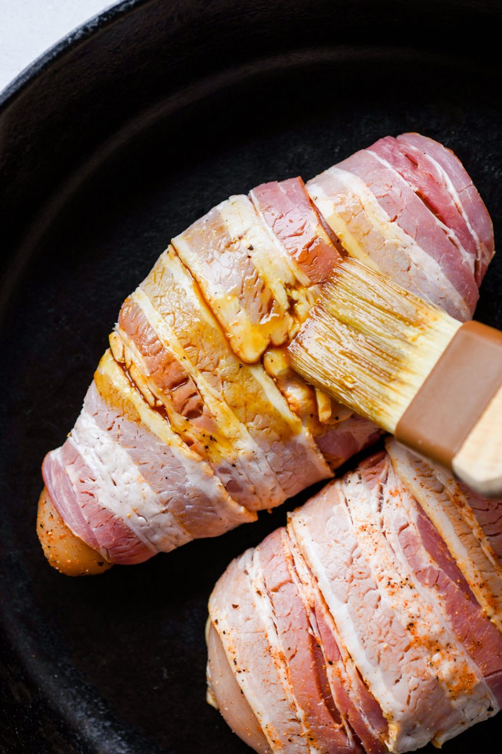 Close up shot of a pastry brush brushing coconut aminos onto a bacon wrapped chicken breast in a cast iron skillet. 