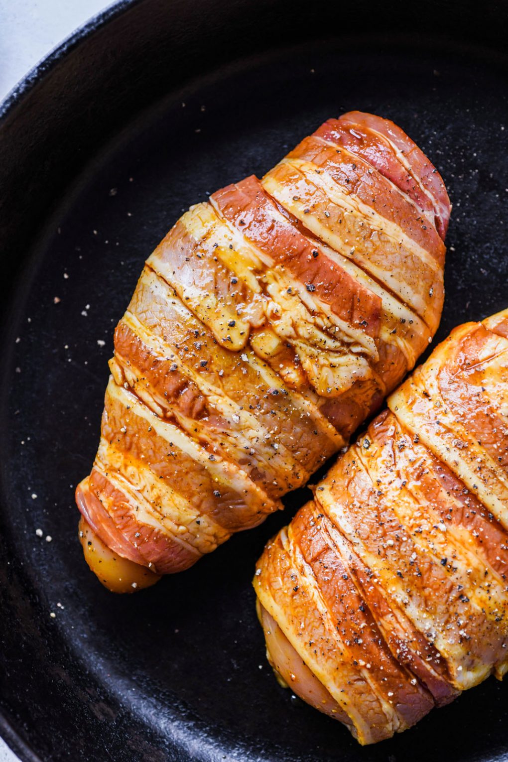 Close up shot of two bacon wrapped chicken breasts in a cast iron skillet, golden brown from being brushed with coconut amino glaze.