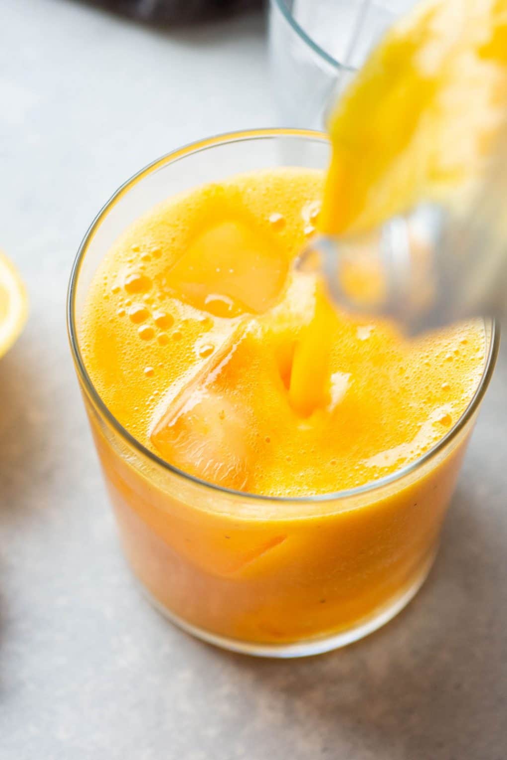 Close up shot of a blender pouring bright orange citrus juice into a cup with ice. On a light grey background. 