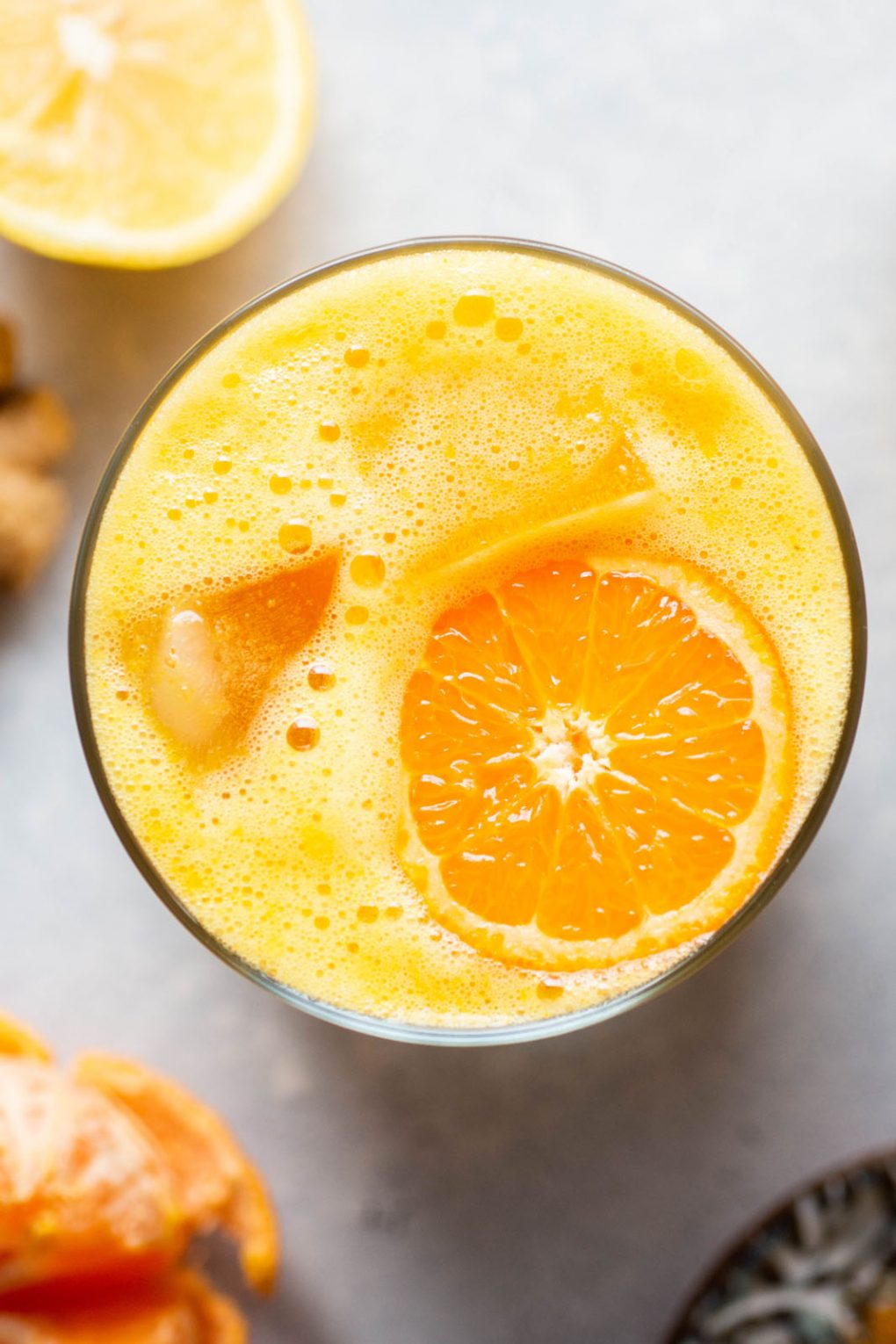 Close up overhead shot of a cup filled with frothy orange Vitamin C citrus juice with a tangerine wheel on top of the juice. On a light grey background next to raw ginger, a half peeled tangerine, and a few slices of lemon. 