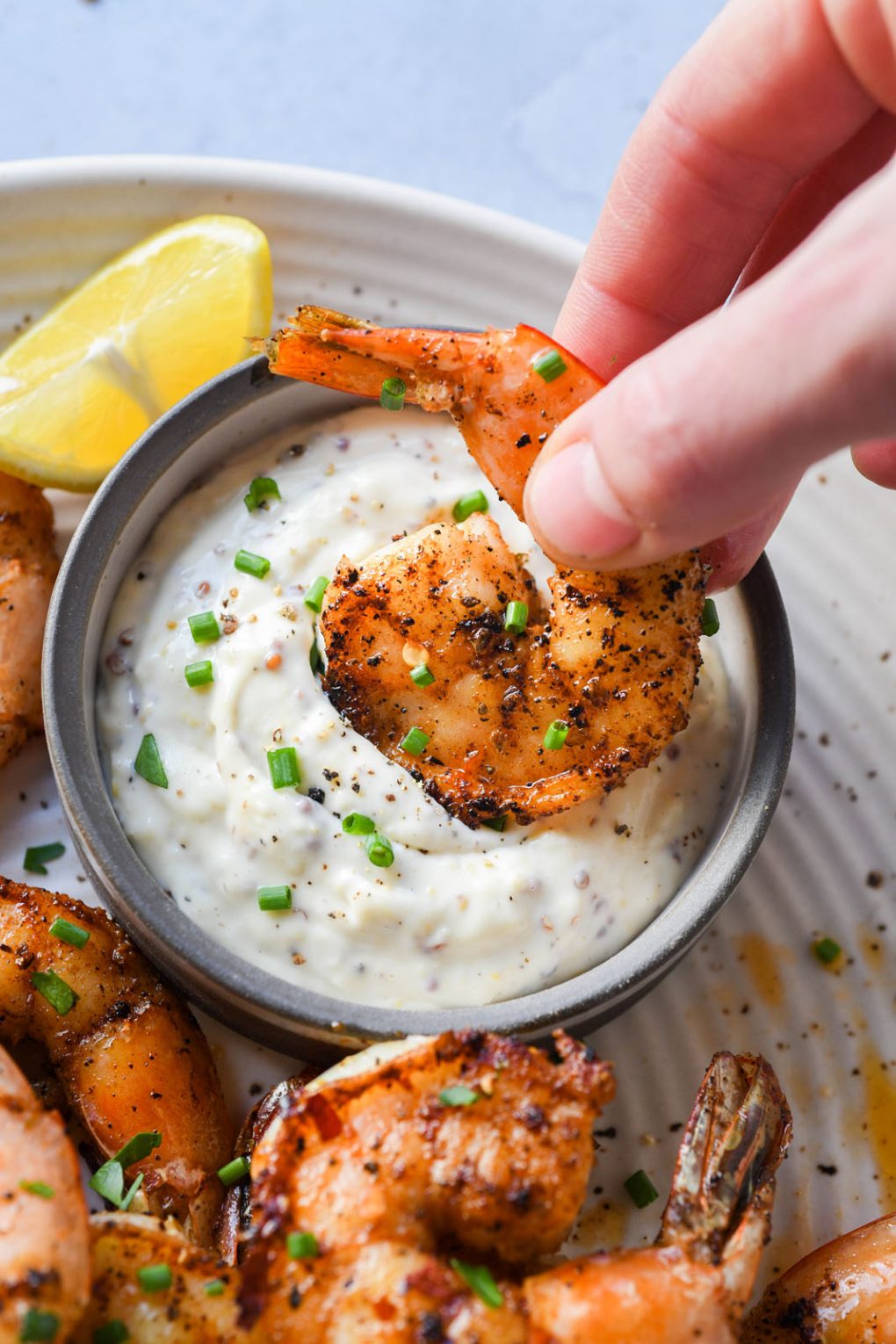 Picture of a hand dipping a cajun spiced shrimp into small bowl of aioli. On a light speckled plate next to a small lemon wedge. 