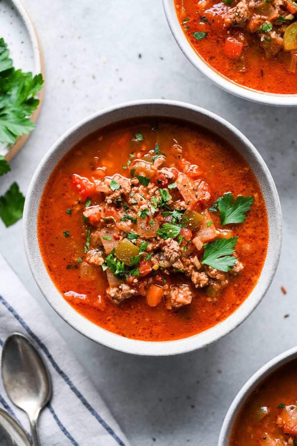 Overhead shot of a colorful bowl of stuffed pepper soup on a light background. On a light background, surrounded by several other bowls of soup and sprigs of fresh parsley. 