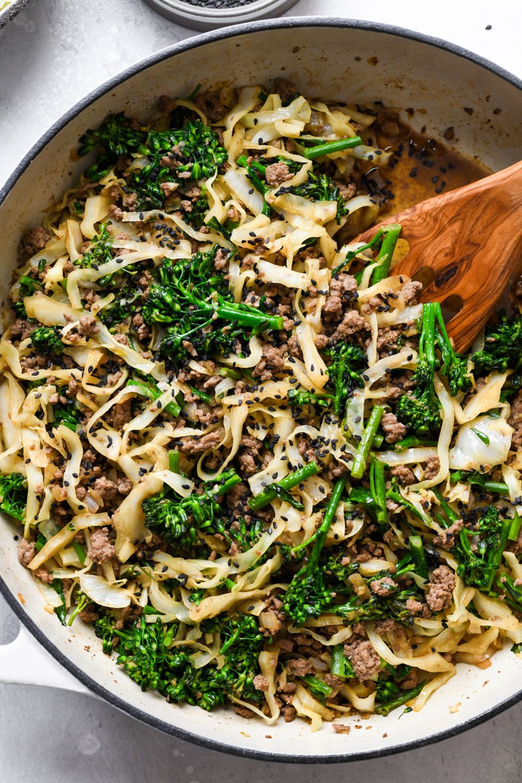 Overhead shot of a large cream colored ceramic skillet filled with ground beef and cabbage whole30 stir fry with cut up broccolini, and topped with black sesame seeds. 